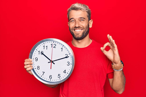 Young handsome blond man with beard doing countdown using big clock over red background doing ok sign with fingers, smiling friendly gesturing excellent symbol - Photo, Image