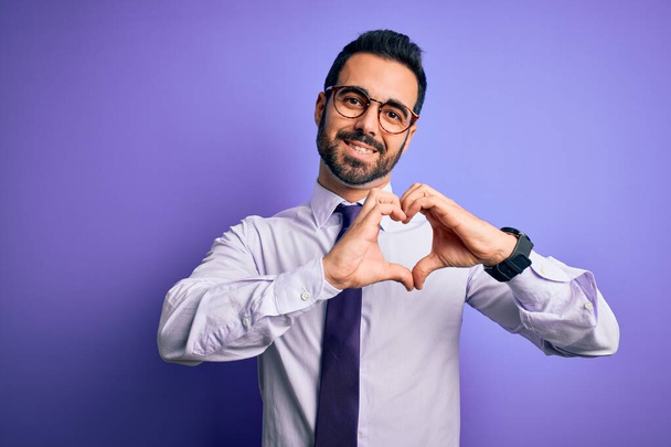 Hombre de negocios guapo con barba con corbata casual y gafas sobre fondo púrpura sonriendo en el amor mostrando el símbolo del corazón y la forma con las manos. Concepto romántico
. - Foto, Imagen