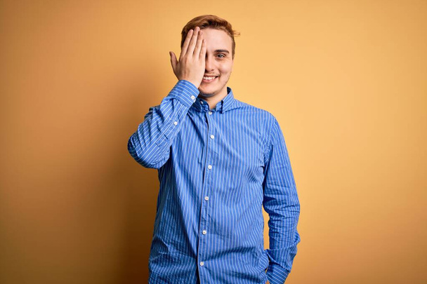 Young handsome redhead man wearing casual striped shirt over isolated yellow background covering one eye with hand, confident smile on face and surprise emotion. - Photo, Image