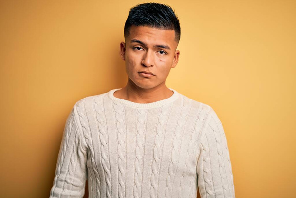 Young handsome latin man wearing white casual sweater over yellow background with serious expression on face. Simple and natural looking at the camera. - Foto, Imagem