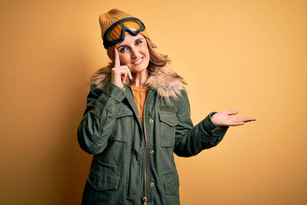 Middelbare leeftijd mooie blonde skiër vrouw dragen sneeuwsportschoenen en skibril verward en geïrriteerd met open palm tonen kopieerruimte en wijzende vinger naar het voorhoofd. Denk er eens over na.. - Foto, afbeelding