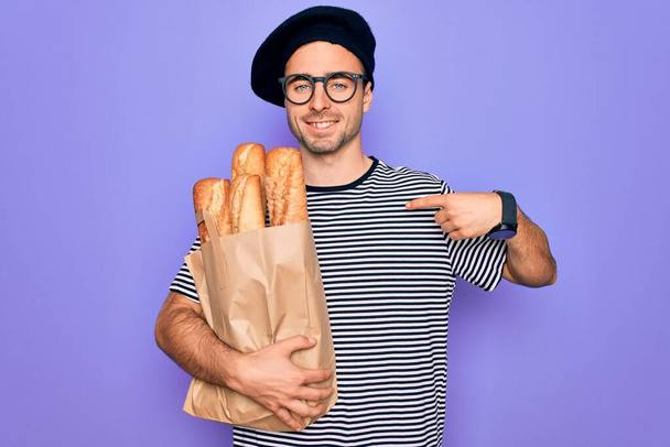 Young handsome baker man with blue eyes wearing french beret holding bag with bread with surprise face pointing finger to himself - Foto, imagen