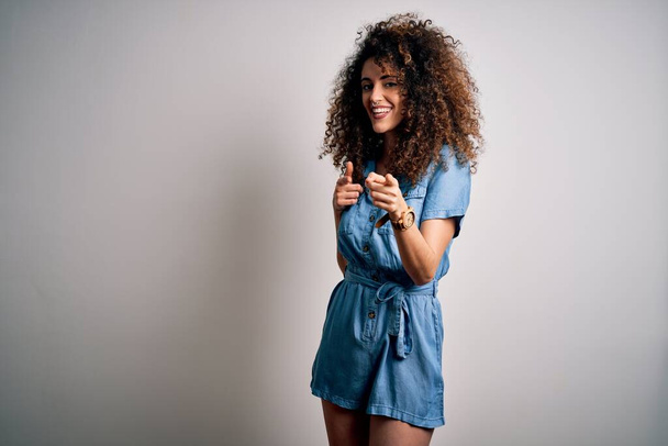 Young beautiful woman with curly hair and piercing wearing casual denim dress pointing fingers to camera with happy and funny face. Good energy and vibes. - Zdjęcie, obraz