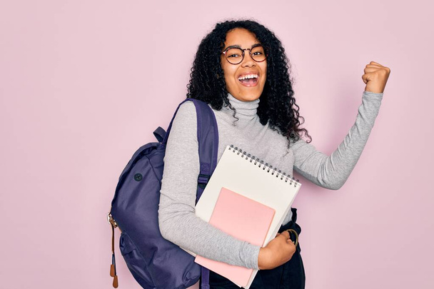 Jovem estudante afro-americana encaracolada usando mochila e óculos segurando livro gritando orgulhoso e celebrando vitória e sucesso muito animado, animando emoção
 - Foto, Imagem