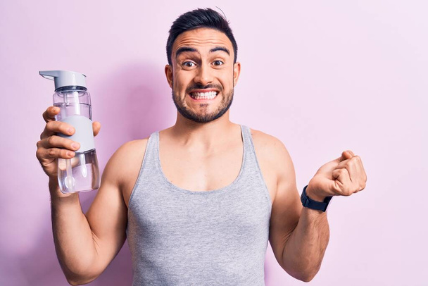 Young handsome sportsman with beard wearing sleeveless t-shirt drinking bottle of water screaming proud, celebrating victory and success very excited with raised arm - Photo, Image