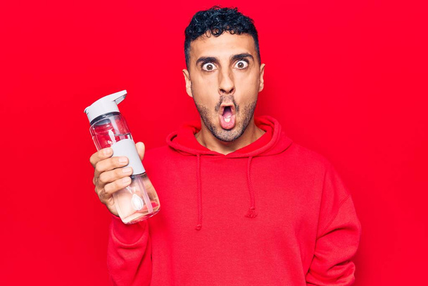 Junger hispanischer Mann trinkt Wasserflasche verängstigt und erstaunt mit offenem Mund für Überraschung, ungläubiges Gesicht  - Foto, Bild