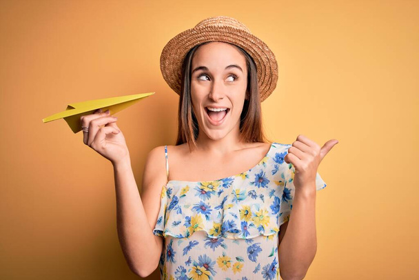 Yaz şapkası takan genç turist kadın sarı arka planda yelpaze tutuyor ve gülümseyen bir gülümsemeyle baş parmağıyla onu işaret ediyor. - Fotoğraf, Görsel