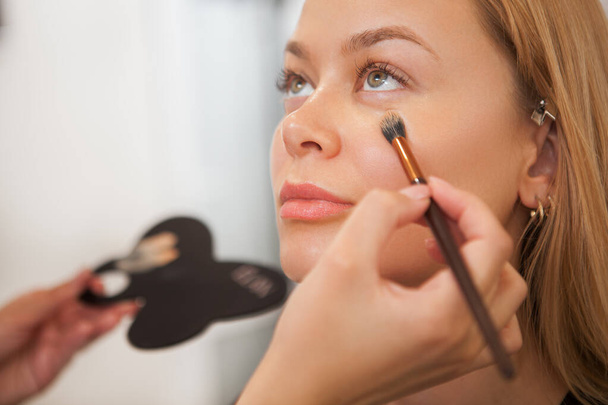 Close up de uma mulher bonita sorrindo enquanto artista de maquiagem profissional aplicando corretor sob seus olhos
 - Foto, Imagem