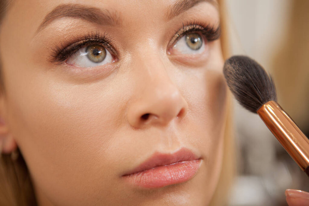 美しい女性の顔にハイライトを適用するプロの化粧アーティストのクロップアップ - 写真・画像
