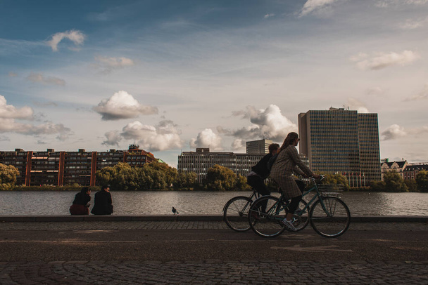 COPENHAGEN, DENMARK - APRIL 30, 2020: People riding bicycles on promenade near canal with buildings and cloudy sky at background  - 写真・画像