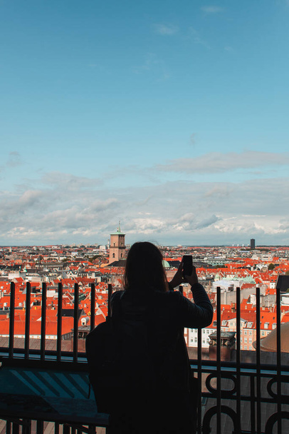 Back view of tourist taking photo on smartphone with buildings and cloudy sky at background in Copenhagen, Denmark  - 写真・画像