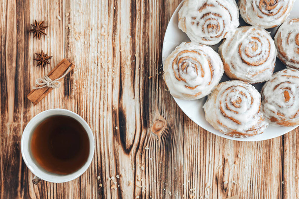 Freshly baked warm iced cinnamon buns on baked paper. Sweet homemade pastry baking on with cup of coffee and spices on wooden background. - Photo, Image