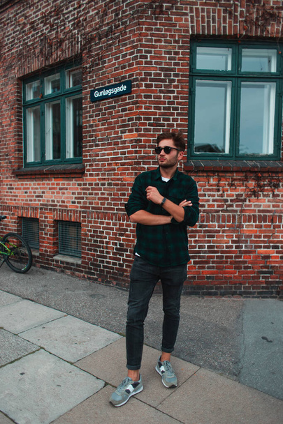 Młody człowiek w okularach przeciwsłonecznych stojący przy fasadzie budynku przy ulicy miejskiej, Kopenhaga, Dania  - Zdjęcie, obraz