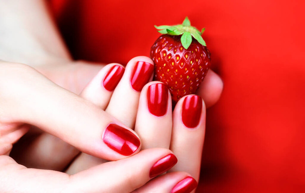 赤い背景にイチゴを持つ赤いマニキュアを持つ女性の手. - 写真・画像