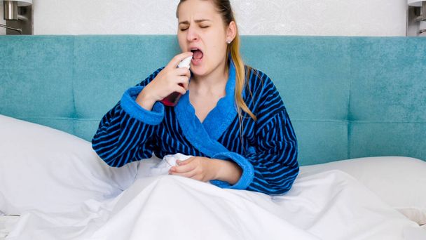 Portrait of young sick woman using throat spray while lying in bed - Photo, Image