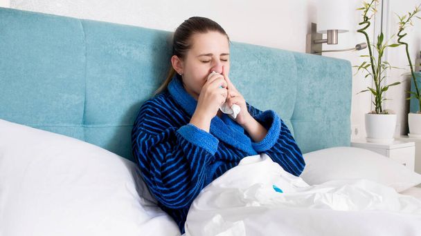 Mujer enferma acostada en la cama sonándose la nariz y usando aerosol nasal
 - Foto, imagen