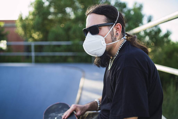 A closeup shot of a bearded male wearing a face mask sitting on a concrete surface while holding his skateboard at the park- Covid-19 concept - Foto, Bild