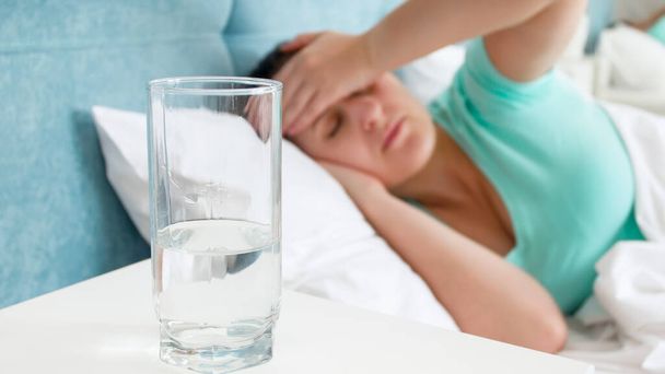 Glass of water on the bedside table against sick woman with migraine lying in bed - Photo, Image