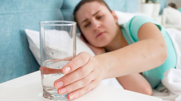 Sick young woman feeling unwell taking glass of water from the bedside table - Photo, Image