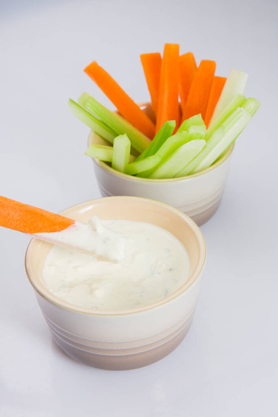close up isolated shot of a bowl of crunchy orange carrot slices and juicy green celery sticks with a single carrot piece dipped in a white cup of blue cheese sauce on a white background - Foto, imagen