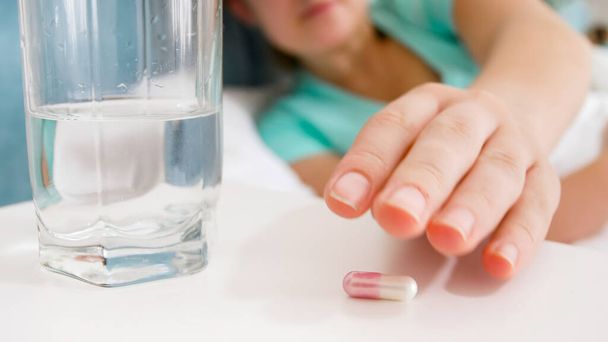 Closeup image of sick woman reaching for pill and glass of water on bedside table - Фото, изображение