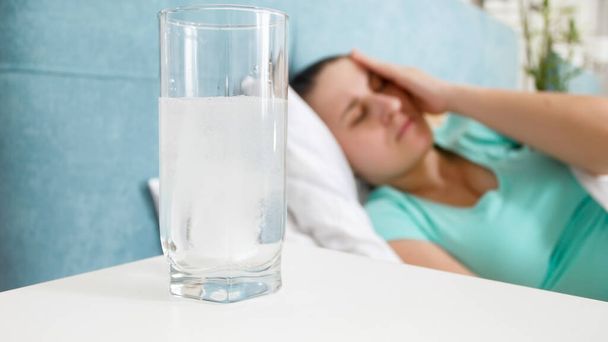 Крупный план стакана аспирина против лежащей в постели женщины, страдающей головной болью
 - Фото, изображение