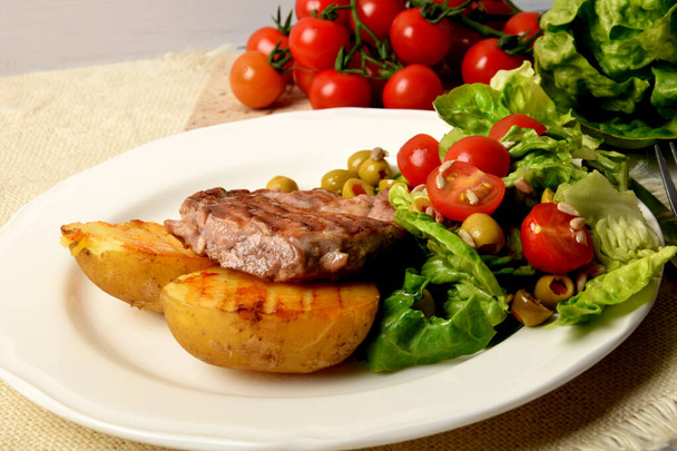 two pieces of grilled potato and grilled meat next to lettuce with tomatoes and olives on a white plate - Foto, Imagem