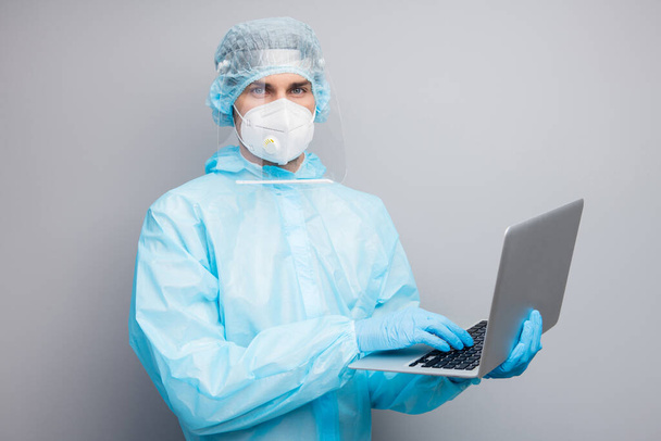 Photo of guy expert doc virology center hold notebook talk patients online call wear respiratory mask hazmat blue uniform suit plastic face protective shield isolated grey color background - 写真・画像