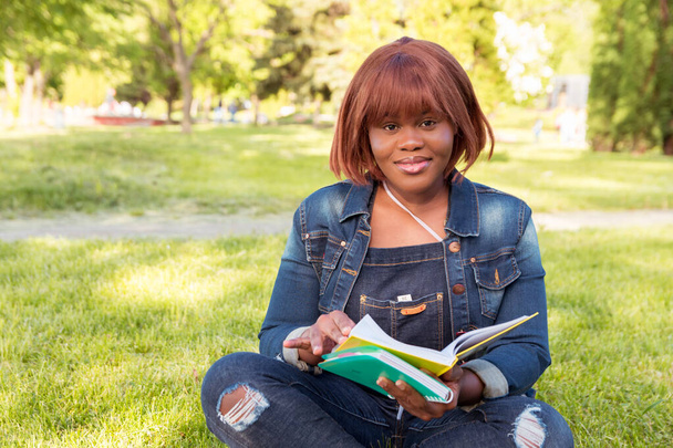 Joven chica negra hermosa sentada en la hierba en el parque sosteniendo cuadernos. Retrato de una estudiante al aire libre
 - Foto, imagen