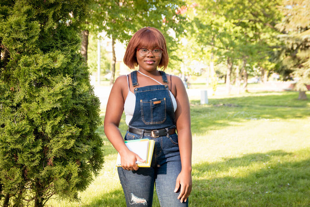 Giovane bella ragazza nera in piedi nel parco con un quaderno in mano. Ritratto di una studentessa all'aperto. Studiare in un'università straniera - Foto, immagini