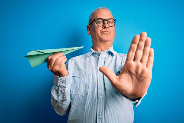 Middelbare leeftijd knappe hamburger man met papieren vliegtuig boven geïsoleerde blauwe achtergrond met open hand doen stop teken met ernstige en zelfverzekerde uitdrukking, verdediging gebaar - Foto, afbeelding