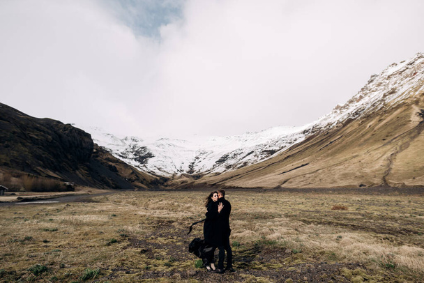Varış yeri İzlanda düğünü. Karlı dağların arka planında bir düğün çifti. Siyah paltolu gelin ve damat yosun ve sarı otların arasında sarılıyorlar.. - Fotoğraf, Görsel