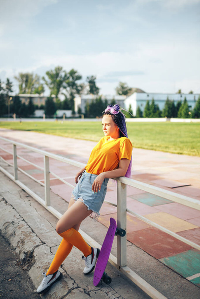 Menina bonita nova inclinada em uma cerca em uma camiseta amarela e meias amarelas, um skate ao ar livre está nas proximidades. - Foto, Imagem