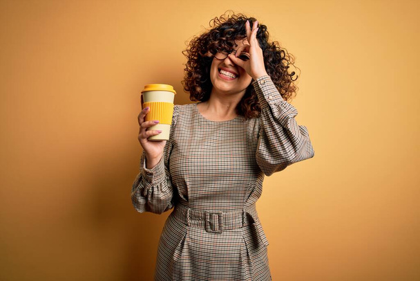 Молодая красивая кудрявая арабская бизнесвумен в очках пьет чашку кофе со счастливым лицом, улыбаясь, делает хорошо знак с рукой на глазу, глядя сквозь пальцы
 - Фото, изображение