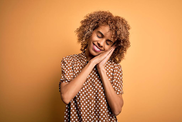 Young beautiful african american woman wearing casual shirt standing over yellow background sleeping tired dreaming and posing with hands together while smiling with closed eyes. - Photo, Image