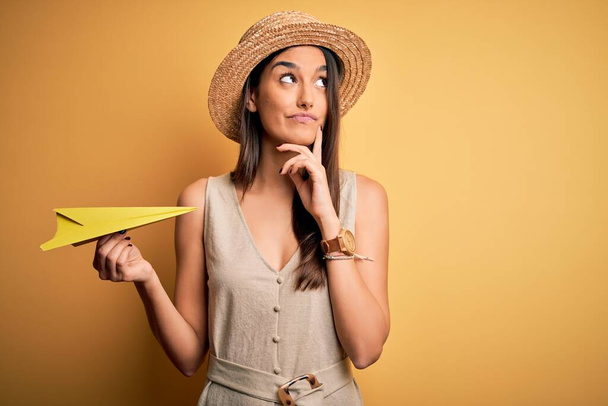 Młoda piękna brunetka kobieta nosi kapelusz trzymając papierowy samolot na żółtym tle poważna twarz myśli o pytaniu, bardzo mylić pomysł - Zdjęcie, obraz