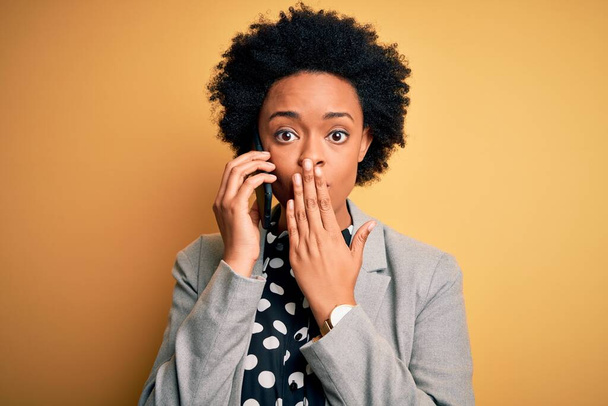 Młoda afro Afroamerykanka z kręconymi włosami rozmawiająca na okładce smartfona z ręką zaszokowana wstydem za pomyłkę, wyrażeniem strachu, przestraszona w ciszy, sekretna koncepcja - Zdjęcie, obraz