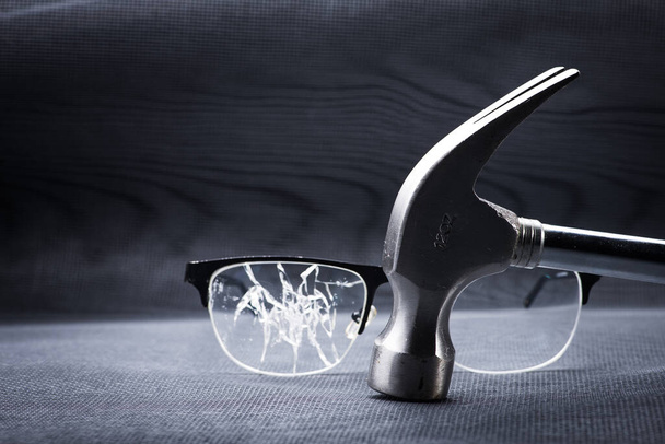 gebroken bril met een metalen hamer op zwarte achtergrond. woede en agressie van een persoon met slecht gezichtsvermogen - Foto, afbeelding