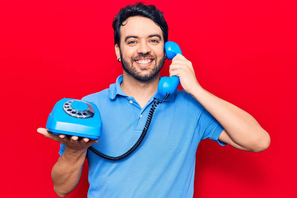 Jovem hispânico segurando telefone vintage olhando positivo e feliz de pé e sorrindo com um sorriso confiante mostrando dentes  - Foto, Imagem