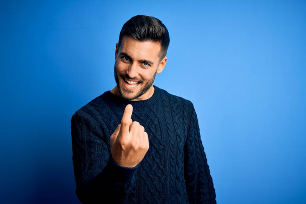 Young handsome man wearing casual sweater standing over isolated blue background Beckoning come here gesture with hand inviting welcoming happy and smiling - Foto, Imagem