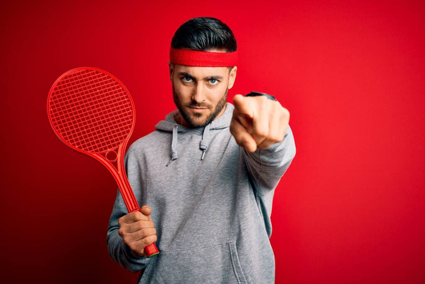 Молодой красивый спортсмен держит теннисную ракетку в спортивной одежде на красном фоне, указывая пальцем на камеру и на вас, знак руки, позитивный и уверенный жест спереди
 - Фото, изображение