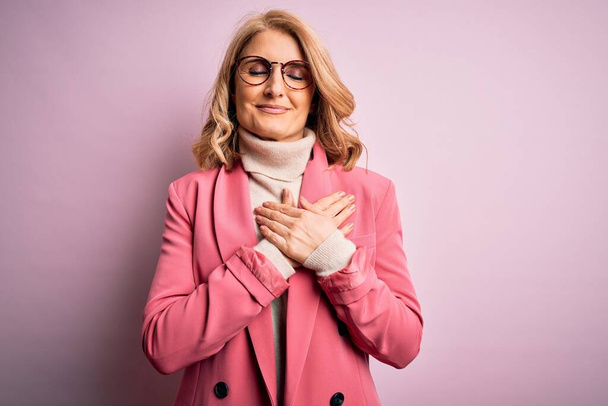 Schöne blonde Geschäftsfrau mittleren Alters in eleganter pinkfarbener Jacke und Brille mit lächelnden Händen auf der Brust, geschlossenen Augen und dankbarer Geste im Gesicht. Gesundheitskonzept. - Foto, Bild