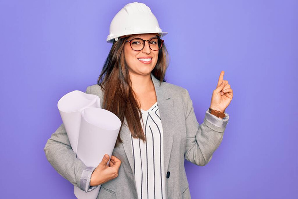 Молодая латиноамериканская архитектор женщина в шляпе строителя и держа чертежи на желтом фоне очень счастливы указывая рукой и пальцем в сторону
 - Фото, изображение