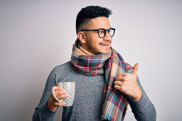 Nuori komea mies yllään huivi juominen muki kuumaa kahvia yli eristetty valkoinen tausta osoittaa ja osoittaa peukalolla sivulle iloinen kasvot hymyillen - Valokuva, kuva