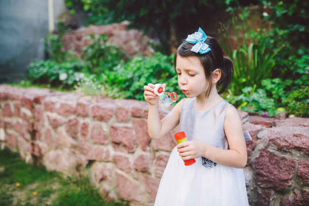 Детские и мыльные пузыри. Девочка развлекается с мыльными пузырями. Маленькая девочка в платье надувает мыльный пузырь в парке. Тема лета и отпуска
. - Фото, изображение