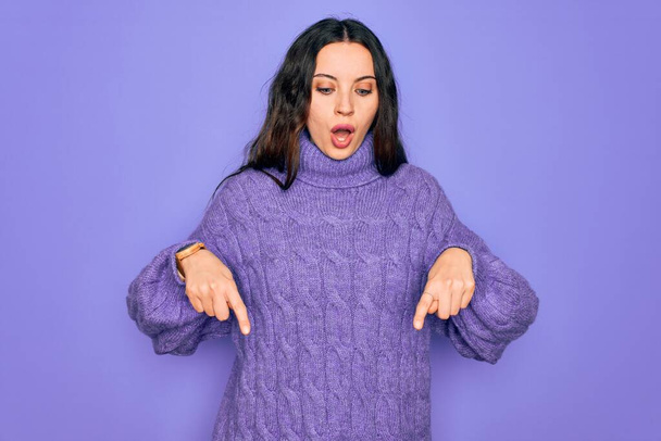 若いです美しいです女性身に着けていますカジュアルturtleeckセーター立って紫色の背景上に指で下に示す広告,驚きの顔とオープン口 - 写真・画像