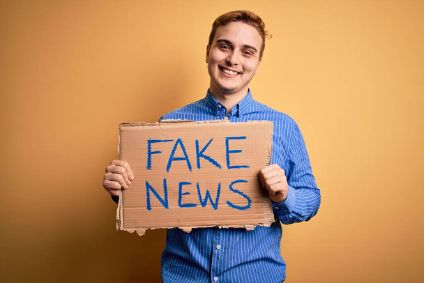 Junger hübscher rothaariger Mann mit einem Banner mit Fake-News-Botschaft auf gelbem Hintergrund, der positiv und glücklich im Stehen aussieht und mit einem selbstbewussten Lächeln lächelt, das Zähne zeigt - Foto, Bild