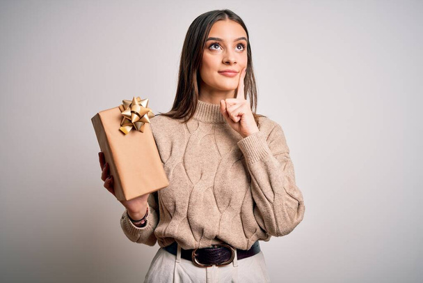 Fiatal gyönyörű barna nő gazdaság születésnapi ajándék felett elszigetelt fehér háttér komoly arc gondolkodás kérdés, nagyon zavaros ötlet - Fotó, kép