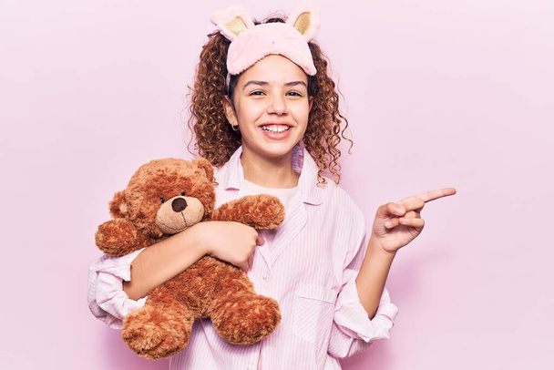 Gyönyörű lány göndör haj visel alvó maszk és pizsama tartó Teddy maci mosolygós boldog rámutatva kéz és ujj az oldalon  - Fotó, kép