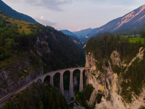 Červený úzkokolejný vlak přes kamenný most na křivolakém úseku trati ve švýcarských Alpách. - Fotografie, Obrázek